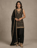 A woman models a traditional black Indian ethnic outfit with intricate gold embroidery, featuring a sleeveless embroidered kurta, black patola-style pants, and a matching black dupatta draped over her shoulder, set against a neutral beige studio background.