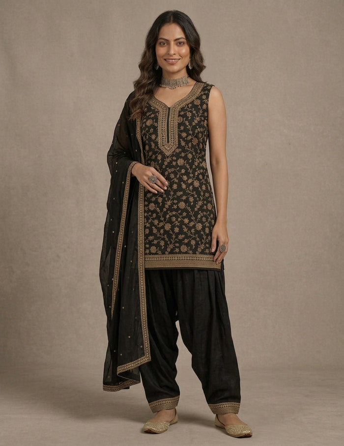 A woman modeling a black traditional outfit with beige floral embroidery, featuring a sleeveless kurta, black pants, and a matching dupatta, set against a neutral beige studio backdrop.