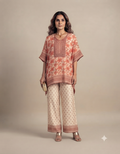 A full-length portrait of a female model against a plain muted beige background, wearing a coral and peach floral-patterned kaftan-style top paired with matching patterned wide-leg trousers, gold heels, a gold necklace, and holding a small gold clutch.