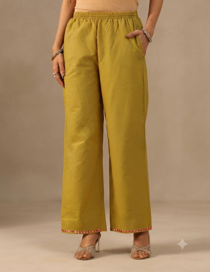 A person modeling wide-leg trousers in a mustard yellow color with a red and gold embroidered border at the hem, paired with beige heeled sandals against a neutral beige studio background.