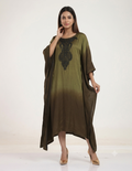 A female model standing against a neutral studio background, wearing an olive-to-brown ombre kaftan dress with intricate black bead embroidery on the neckline, paired with gold heels.