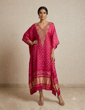 A woman models a bright pink kaftan dress with gold embroidery, white polka dots, and a gold border, standing against a textured beige wall.