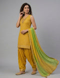 A woman modeling a traditional Indian salwar kameez suit in mustard yellow with a green and yellow gradient dupatta, set against a plain gray studio background.